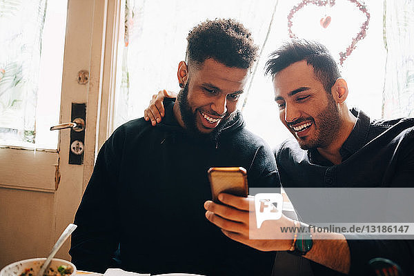 Smiling young men sharing smart phone while sitting against window at restaurant during brunch party