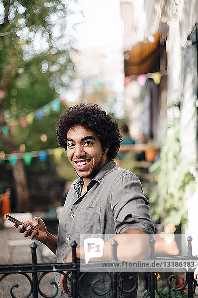 Porträt eines lächelnden jungen Mannes  der sein Mobiltelefon in der Hand hält  während er sich im Hinterhof an ein Geländer lehnt