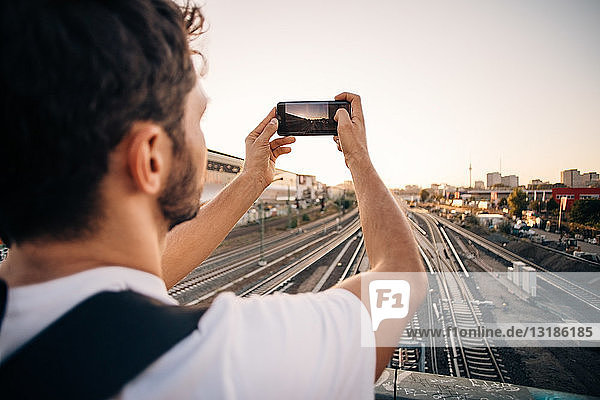 Young man photographing through mobile phone over railroad tracks in city