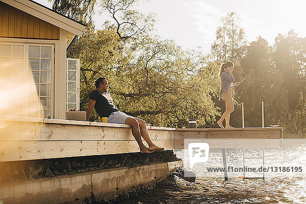 Erwachsener Mann sieht Frau mit digitalem Tablett an  während er auf der Terrasse am See sitzt