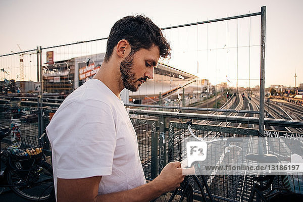 Side view of young man using mobile phone while standing on bridge over railroad tracks in city