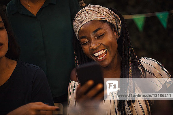 Young woman laughing while looking at mobile phone during dinner party with friends in backyard