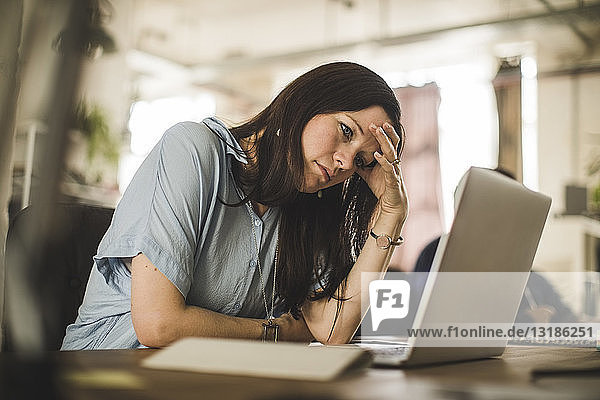 Müde Geschäftsfrau schaut auf den Laptop  während sie im Kreativbüro am Schreibtisch sitzt