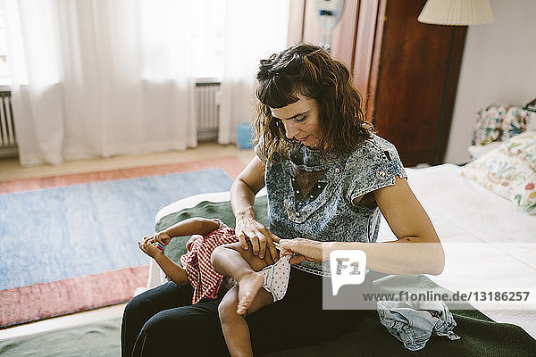 Mother adjusting diaper of daughter while sitting on bed at home
