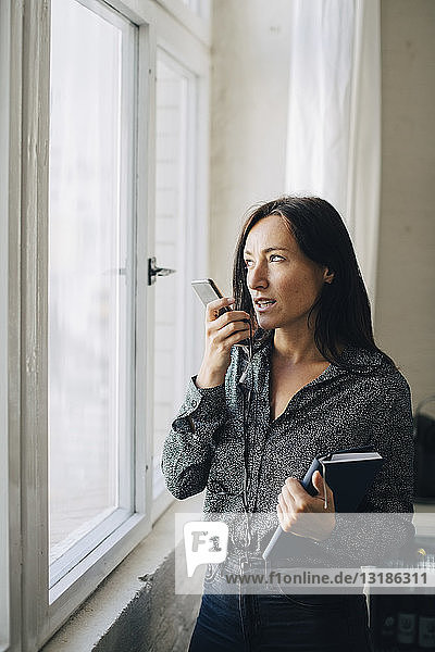 Kreative Geschäftsfrau spricht mit dem Handy  während sie im Büro am Fenster steht