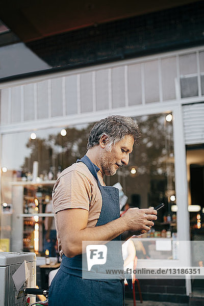 Side view of male owner using mobile phone outside deli