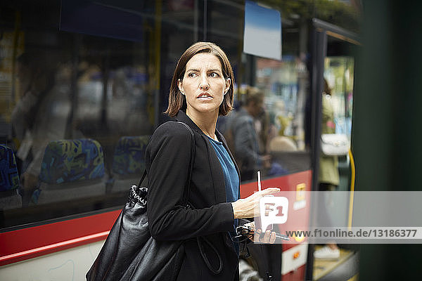 Confident businesswoman looking away while standing with drink against bus in city