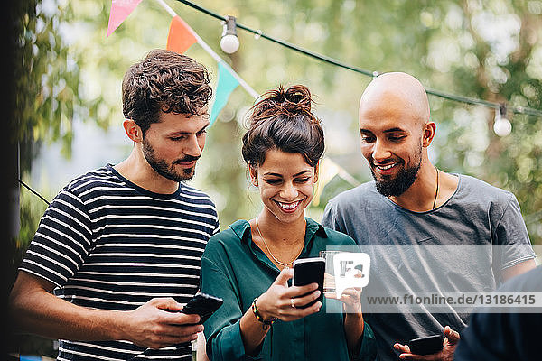Glückliche junge Frau zeigt männlichen Freunden ihr Handy  während sie während einer Party auf dem Balkon steht