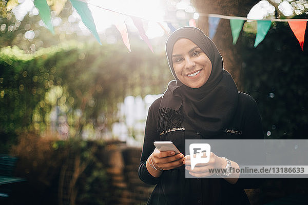 Porträt einer lächelnden Frau im Hijab mit einem Smartphone in der Hand im Hinterhof