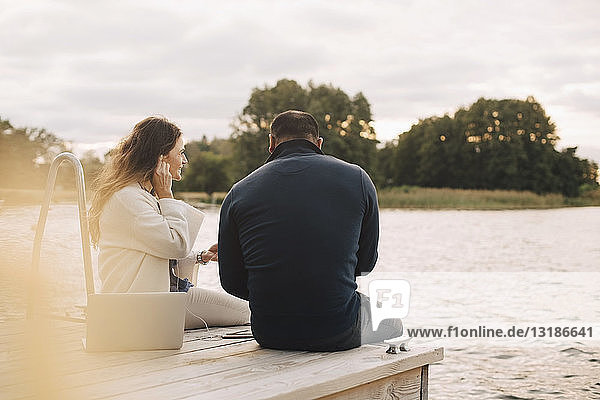 Rückansicht eines Paares  das mit einem Smartphone Musik hört  während es auf einem Steg am See gegen den Himmel sitzt
