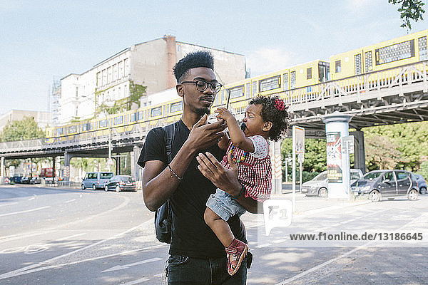 Young man carrying daughter holding mobile phone while standing on street against railway bridge