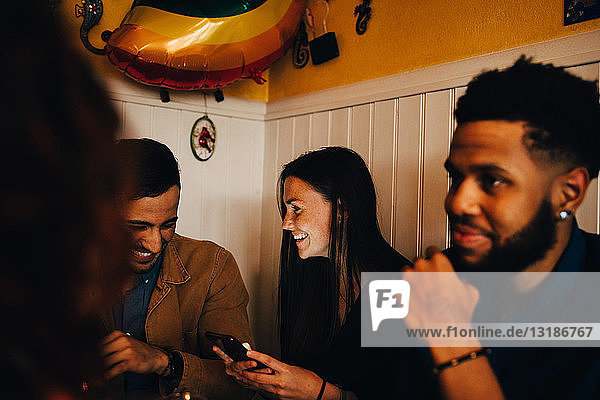 Junge Frau und Mann teilen sich ein Mobiltelefon  während sie bei einem Freund im Restaurant sitzen