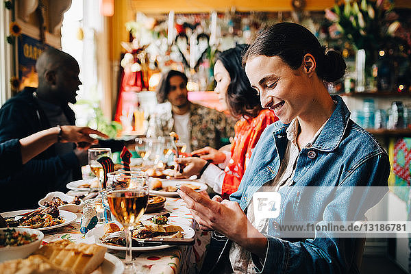 Lächelnde junge Frau  die ein Smartphone benutzt  während sie während einer Dinnerparty mit Freunden im Restaurant am Tisch sitzt