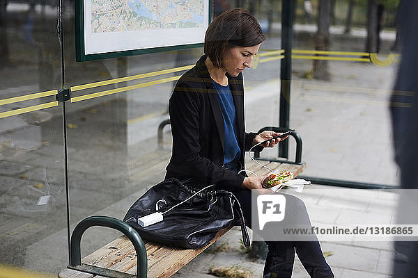 Mittlere erwachsene Geschäftsfrau benutzt ein Smartphone  während sie an einer Bushaltestelle in der Stadt ein Sandwich isst