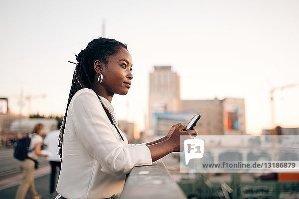 Nachdenkliche junge Frau schaut weg  während sie ihr Handy auf der Brücke in der Stadt in der Hand hält