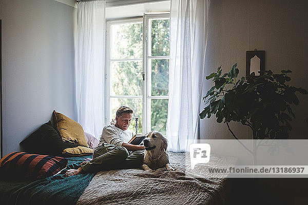 Full length of retired man reading book while resting with dog on bed at home