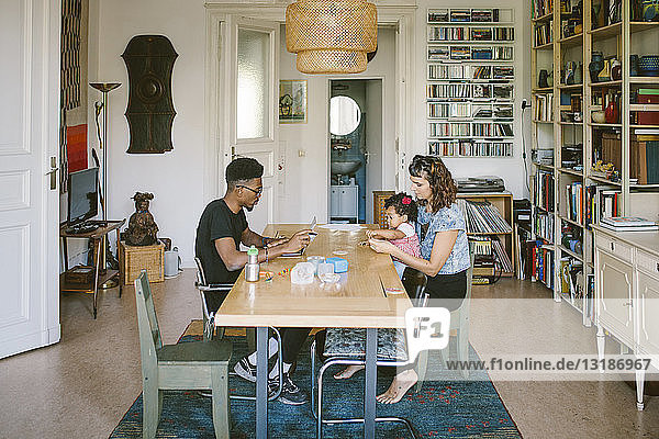 Junger Mann benutzt Laptop  während Frau mit Tochter am Esstisch im Haus spielt