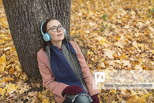 Gelassene Frau  die mit Kopfhörern unter einem Baum im herbstlichen Park Musik hört