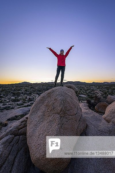 Junge Frau steht mit ausgestreckten Armen auf runden Granitfelsen bei Sonnenuntergang  Felsformationen  White Tank Campground  Joshua Tree National Park  Desert Center  Kalifornien  USA  Nordamerika