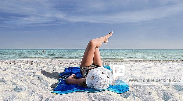 Mädchen mit Sonnenhut auf einem blauen Strandtuch am Meer an einem weißen Sandstrand liegend