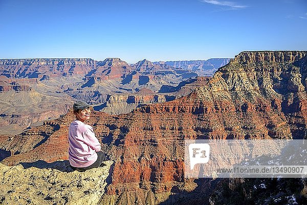 Junge lächelnde Frau  sitzend am Abgrund der gigantischen Schlucht des Grand Canyon  Blick vom Rim Trail  zwischen Mather Point und Yavapai Point  erodierte Felslandschaft  South Rim  Grand Canyon National Park  nahe Tusayan  Arizona  USA  Nordamerika