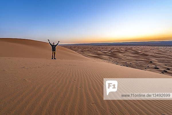 Young man stretches his arms into the air  sand dune at sunrise  Erg Chebbi  Merzouga  Sahara  Morocco  Africa