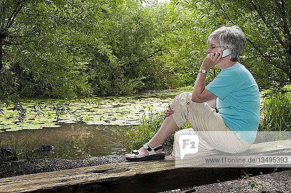 Frau sitzt auf einer Bank an einem Waldsee und telefoniert mit ihrem Mobiltelefon