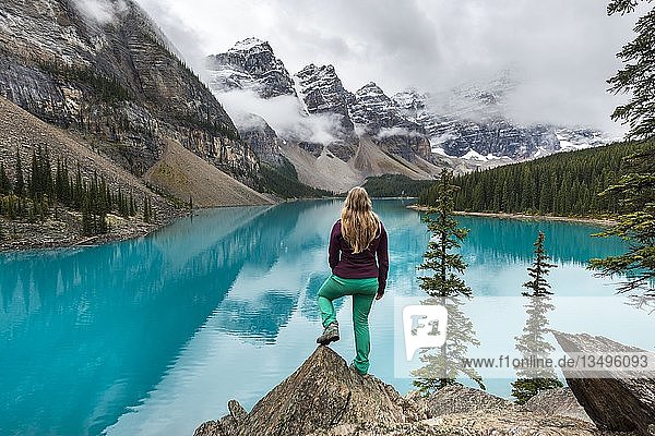 Young woman standing in front of a lake looking into mountain scenery  clouds hanging between mountain peaks  reflection in turquoise lake  Moraine Lake  Valley of the Ten Peaks  Rocky Mountains  Banff National Park  Province of Alberta  Canada  North America