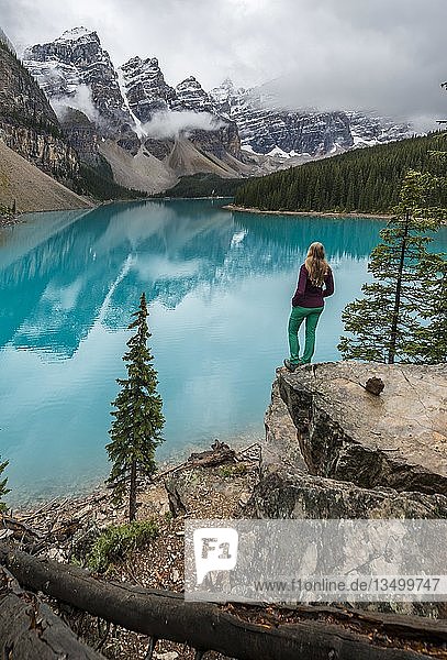 Junge Frau steht vor einem See und blickt in eine Berglandschaft  Wolken hängen zwischen Berggipfeln  Spiegelung im türkisfarbenen See  Moraine Lake  Valley of the Ten Peaks  Rocky Mountains  Banff National Park  Provinz Alberta  Kanada  Nordamerika