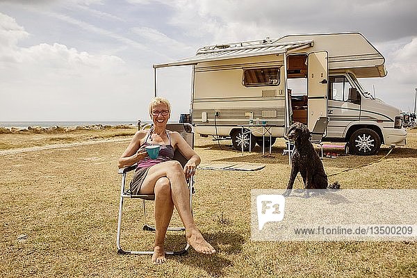 Frau sitzt mit einem Hund  Königspudel  vor ihrem Wohnmobil und trinkt Kaffee während eines Urlaubs am Meer  Portbail  Normandie  Frankreich  Europa