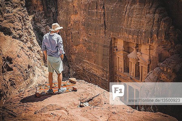 Tourist mit Sonnenhut steht auf Felsen und schaut von oben in die Schlucht Siq  in den Fels gehauenes Schatzhaus des Pharao  Fassade des Schatzhauses Al-Khazneh  Khazne Faraun  Mausoleum in der Nabatäerstadt Petra  nahe Wadi Musa  Jordanien  Asien