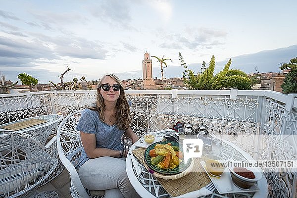 Junge Frau auf einer Dachterrasse im Restaurant marokkanisches Essen Tajine Ausblick auf die Altstadt Moschee mit Minaret Abendstimmung Marrakech Marokko Afrika