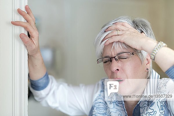 Ältere Frau mit Kopfschmerzen hält sich die Hand auf die Stirn und stützt sich auf den Türrahmen  Deutschland  Europa