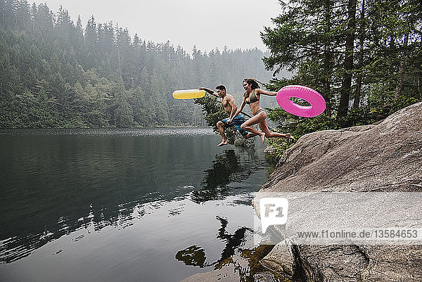 Verspieltes junges Paar mit aufblasbaren Ringen  das in einen abgelegenen See springt  Squamish  British Columbia  Kanada