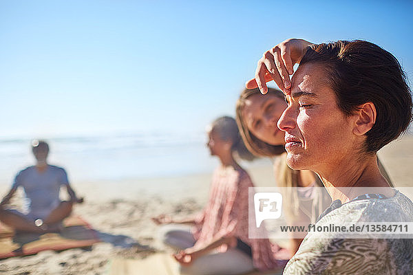 Yoga instructor touching third eye of woman meditating on sunny beach during yoga retreat