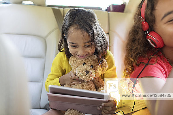 Niedliches Mädchen mit Teddybär  das ein digitales Tablet auf dem Rücksitz eines Autos benutzt