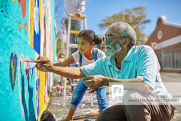 Grandfather and granddaughter volunteers painting vibrant mural on sunny urban wall