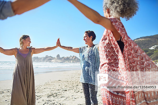 Women joining hands in circle on sunny beach during yoga retreat