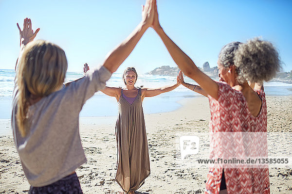 Women joining hands in circle on sunny beach during yoga retreat
