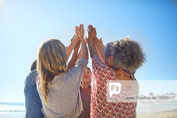 Freundinnen erheben die Hände im Kreis während eines Yoga-Retreats am sonnigen Strand