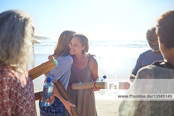 Freundinnen mit Yogamatten umarmen sich am sonnigen Strand während eines Yoga-Retreats