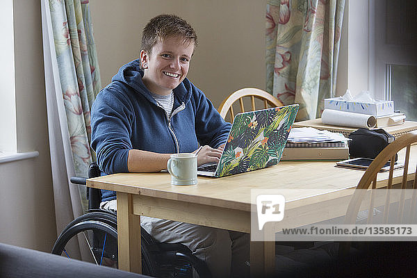 Porträt einer lächelnden  selbstbewussten jungen Frau im Rollstuhl  die einen Laptop am Esstisch benutzt