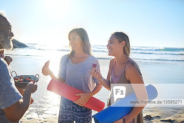 Mutter und Tochter mit Yogamatten essen frische Beeren am sonnigen Strand während eines Yoga-Retreats
