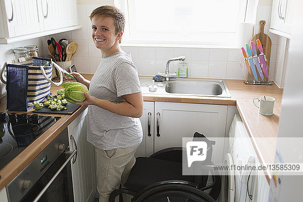 Porträt einer selbstbewussten jungen Frau im Rollstuhl  die in einer Wohnküche kocht