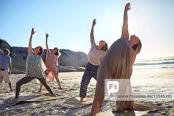 Group practicing yoga reverse warrior pose on sunny beach during yoga retreat