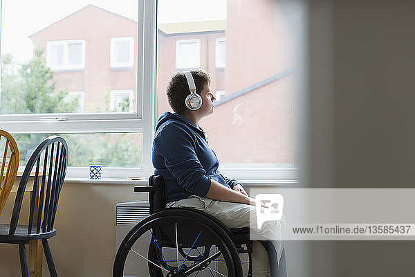 Nachdenkliche junge Frau im Rollstuhl  die mit Kopfhörern am Fenster Musik hört