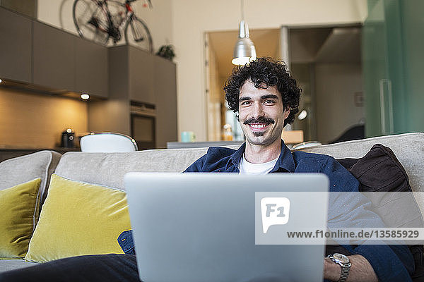 Porträt eines lächelnden Mannes mit Laptop auf dem Sofa einer Wohnung