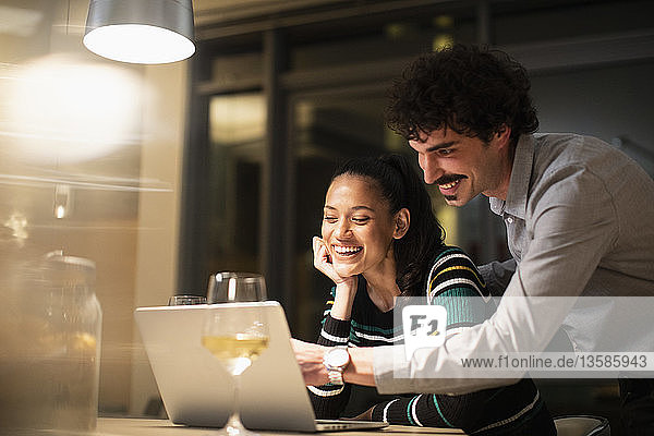 Glückliches Paar  das abends zu Hause einen Laptop benutzt und Weißwein trinkt