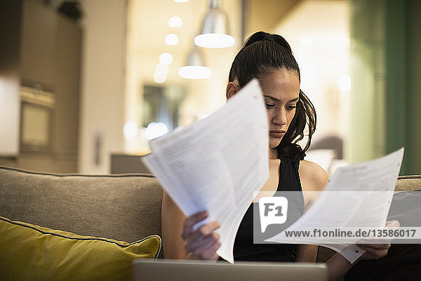 Woman reading paperwork at laptop on sofa