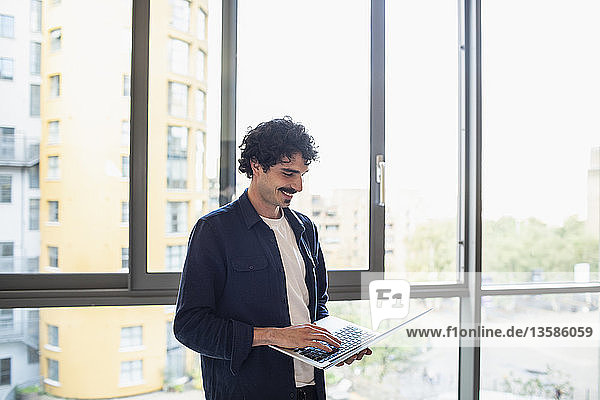 Lächelnder Mann mit Laptop am Fenster einer städtischen Wohnung
